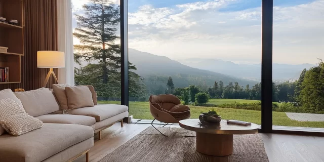 A modern living room, potentially boosting home value through renovation, boasts a comfortable beige sectional sofa and a cozy chair around a round wooden table on a woven rug. Floor-to-ceiling windows reveal serene mountains and trees under a bright, partly cloudy sky.