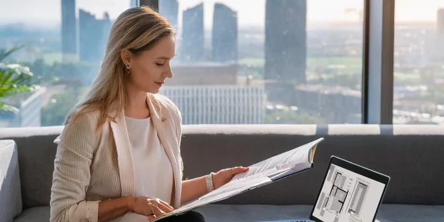 A woman sits on a sofa in a modern office, engrossed in a magazine about condo renovations. Beside her, a laptop rests ready for research. The large windows reveal a city skyline with skyscrapers, while natural light floods the room.