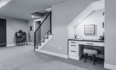 a modern basement features a staircase with sleek railings, a desk with a stool under an angled ceiling, and a motivational quote on the wall. the minimalist design is accented by a small seating area in the background.