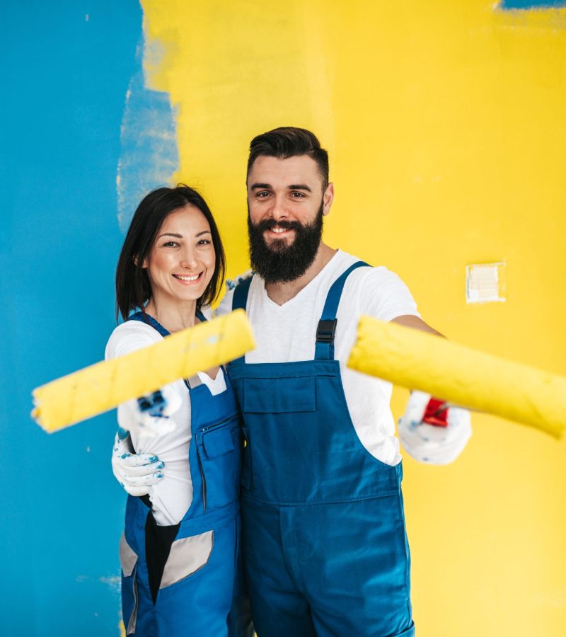 a smiling couple wearing blue overalls holds paint rollers in front of a half-painted wall. the wall is painted in two bold colors: blue on the left and yellow on the right. they appear to be in the middle of a painting project.