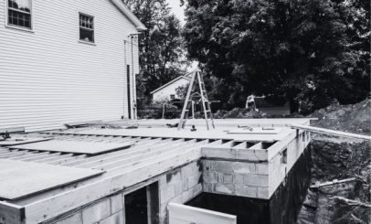 a monochrome image of a house under construction. wooden beams form the frame for an annex. a ladder stands on the structure, with various tools scattered around. trees and more houses are visible in the background.