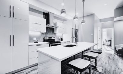 a modern kitchen with a sleek design features a long island counter with three barstools. pendant lights hang above the island. stainless steel appliances, including an oven and fridge, are set against white cabinetry and walls.