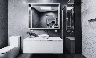 a modern bathroom in black and white, featuring a sleek white sink on a marble countertop, a large mirror above, a toilet to the left, and a glass-enclosed shower to the right. the walls are tiled in a herringbone pattern.