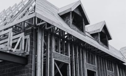 black and white photo of a house under construction. the skeletal wooden framework is visible, with roof trusses and dormer windows in progress. the structure is set against a plain sky.
